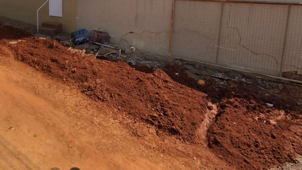 Terra de obra municipal acumulada junto a muro de residência com rachaduras, exemplo de dano causado por obra da prefeitura